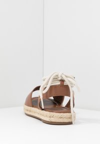 Sandale en cuir marron avec lien en corde blanche, dos ouvert, semelle en jute, et design plat, comportant des coutures minimales et des accents métalliques.