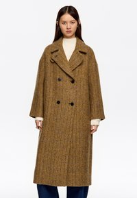 Abrigo de lana de doble botonadura con patrón de espiga, que presenta dos botones negros, solapas grandes y mangas anchas en tonos marrón y amarillo.