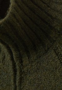 Suéter de punto en verde oscuro, con un cuello alto acanalado y una superficie texturada y difusa. Primer plano del patrón detallado de la tela.