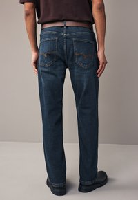 Granatowe dżinsy o prostym kroju, z klasycznymi tylnymi kieszeniami ozdobionymi pomarańczowymi przeszyciami. Noszone z czarnymi butami.