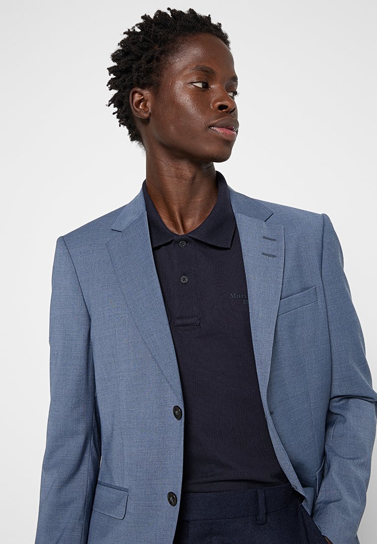 Junger Mann mit kurzen lockigen Haaren, der einen hellblauen Blazer über einem dunkelblauen Poloshirt trägt und zur Seite schaut, vor einem einfarbigen Hintergrund.