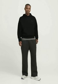 Hombre de pie que lleva una sudadera negra, pantalones gris oscuro, camiseta interior gris y zapatillas blancas frente a un fondo claro y liso.