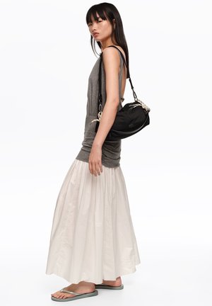 Mujer con top gris sin mangas, falda larga blanca, chanclas grises, llevando un bolso de hombro negro con detalles de cuerda.