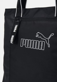 Sac cabas en nylon noir avec logo Puma blanc, fermeture éclair et anses rayées contrastantes. Surface texturée avec motifs géométriques.