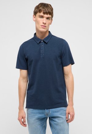 Junger Mann trägt ein dunkelblaues kurzärmeliges Poloshirt und hellblaue Jeans, steht vor einem einfarbigen hellen Hintergrund.