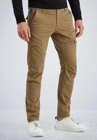 Man in kaki cargo shorts en witte sneakers, staand met ontspannen handen langs zijn zij tegen een eenvoudige achtergrond.