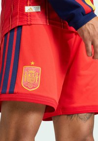 Espanjan maajoukkueen punaiset polyesterishortsit, joissa on kolme tummansinistä raitaa, teksturoitu logopatchi kruunulla ja keltaiset yksityiskohdat.