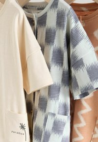 Tri kratke majice s kratkimi rokavi na obešalniku: kremne barve z napisom »paradise« in motivom palme, bele z sivo karo vzorcem ter oranžne z vzorcem cik-cak.