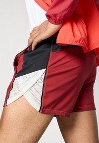 Κόκκινα και μαύρα αθλητικά σορτς με λευκές μεσαίες λεπτομέρειες, διαθέτουν πλαϊνές σχισμές, υφασματική υφή και αντίθετες ραφές.