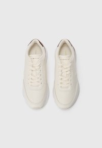 Une paire de sneakers décontractées off-white avec des lacets, des accents de talon marron et un logo subtil, vue de dessus sur un fond blanc.