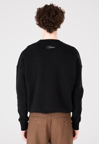 Person mit lockigem Haar, der einen schwarzen Pullover mit dem Schriftzug "Verboten" auf dem oberen Rücken und braune Hosen trägt und von der Kamera wegsteht.