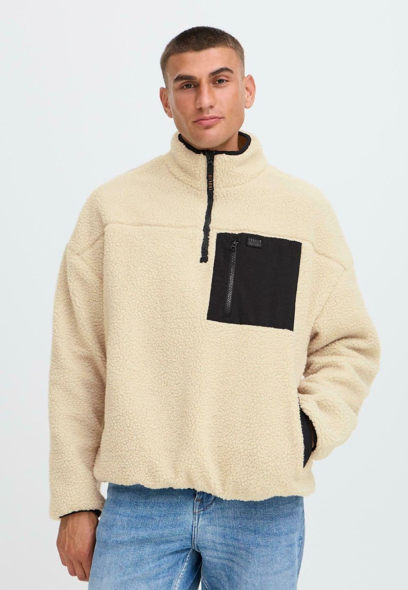 Suéter de forro polar en color crema con un bolsillo negro en el pecho, con un cuello alto y cierre de media cremallera. Textura suave y corte relajado.