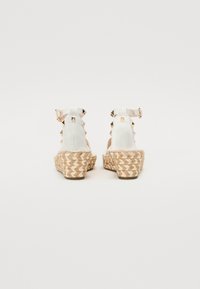 Sandales compensées blanches avec des semelles tressées texturées, ornées de clous en or rose le long des sangles de talon et de boucles de cheville ajustables.
