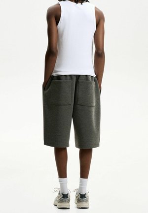 Persoon staand met de rug naar voren, gekleed in een wit mouwloos shirt, donkergrijze knielange shorts met achterzakken, witte sokken en beige sneakers.