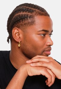 Orecchino a forma di diamante in oro appeso all'orecchio sinistro, abbinato a capelli scuri intrecciati e una maglietta nera. Mani incrociate sotto il mento.