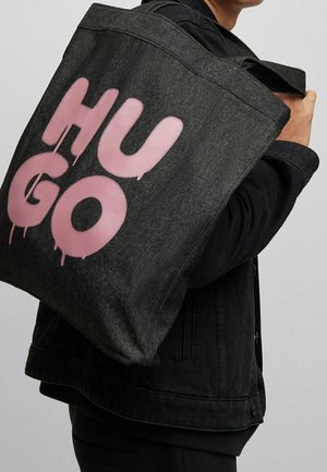Personne portant une veste en denim noire avec un grand sac fourre-tout noir affichant un texte audacieux "HUGO" en style graffiti rose vif.