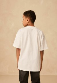Tricou alb din bumbac, cu mâneci scurte și decolteu rotund, având o croială lejeră. Fundalul este de o nuanță neutră delicată.
