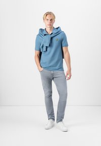 Lys blå polo-skjorte med en brettet hette, gul logo, kombinert med lysegrå jeans og hvite joggesko. Enkel, uformell design.