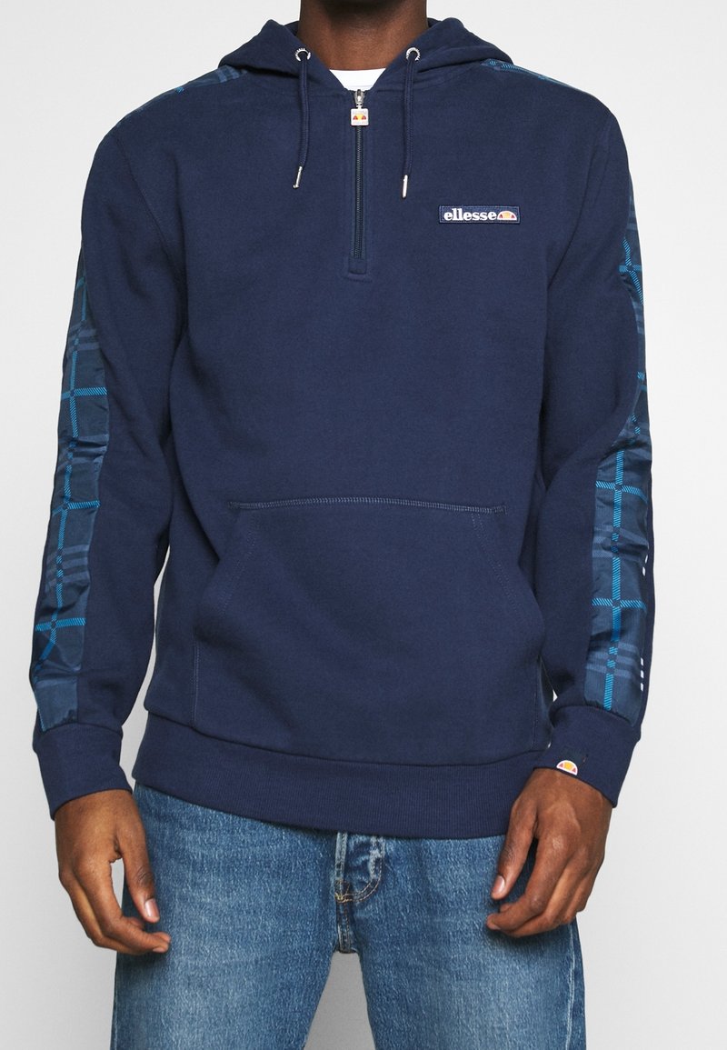 Homme portant un sweat à capuche bleu marine ellesse avec demi-fermeture éclair et motif à carreaux sur les manches, associé à un jean bleu sur fond blanc.