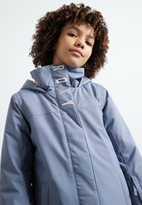 Casaco impermeável azul claro com gola alta, lado com a marca "ROXY" e fecho frontal. Textura suave e design desportivo.