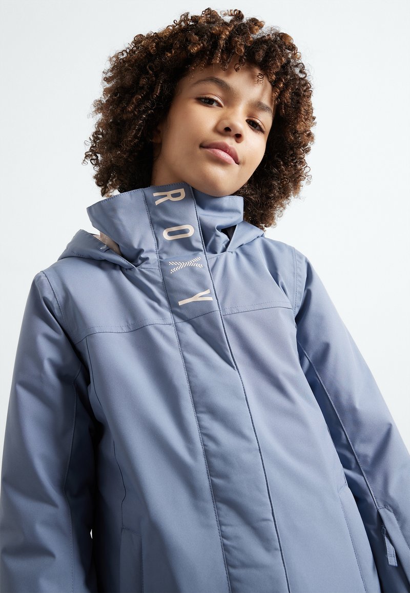 Casaco impermeável azul claro com gola alta, lado com a marca "ROXY" e fecho frontal. Textura suave e design desportivo.