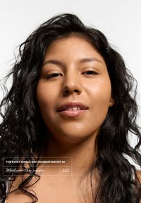 Tubo de base con un tono cálido medio-moreno 625, destacado con la etiqueta "SPF 50". Modelo con cabello negro ondulado, fondo neutro.