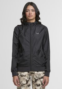 Black zip-up jacket in lightweight fabric with a hood, elastic cuffs, and waistband. Features subtle butterfly logo on the chest.