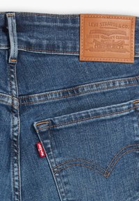Jeans de mezclilla azul con un parche de cuero de la marca. Presentan costuras naranjas y una etiqueta de logo roja en el bolsillo trasero. Detalles de diseño remachados.