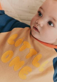 Langærmet shirt i beige med orange detaljer. Har afrundede gule bogstaver, der staver "LITTLE ONE" på fronten. Blød tekstur.