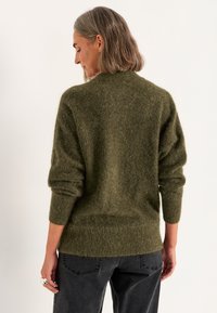 Olivengrüner Wollpullover mit lockerer Passform, gerippten Bündchen und einer weichen, strukturierten Oberfläche, getragen über dunkelgrauen Hosen.