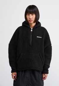 Sweat à capuche en polaire noire avec une fermeture éclair à demi, grande poche avant et détail de logo sur la poitrine. Texture douce et coupe oversize.