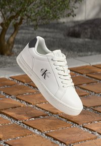 Baltos sportinės kedai su lygiu odai panašiu medžiaga, su raišteliais, pilku kulno akcentu ir iškiliu logotipu „Calvin Klein Jeans“ ant šono.