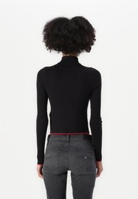 Černý žebrovaný pletený svetr s rolákem, přiléhavého střihu a dlouhými rukávy; červený akcent na spodním lemu, nošený s šedými džíny.