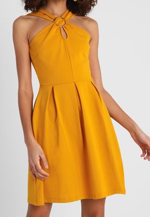 Femme portant une robe sans manches jaune moutarde avec un col halter et un détail anneau en métal au niveau de la poitrine, debout devant un fond uni.