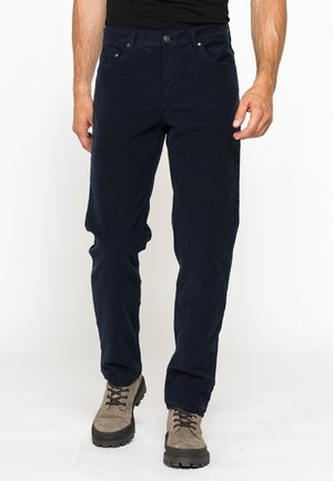 Uomo con pantaloni blu scuro e scarponi da trekking beige, in piedi davanti a uno sfondo bianco uniforme.