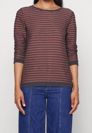 Donna che indossa una camicia a maniche lunghe a righe orizzontali marroni e nere e jeans blu, in piedi su uno sfondo neutro.