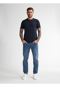 Granatowa koszulka z krótkim rękawem, okrągły dekolt, zestawiona z jasnoniebieskimi dżinsami i białymi trampkami. Zawiera widoczne tatuaże na ramionach.