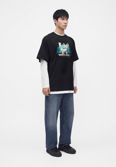 Νέος άνδρας φοράει μαύρο φαρδύ t-shirt με "Rebel" και γραφικό σκύλου πάνω από λευκή μακρυμάνικη μπλούζα, μπλε τζιν και μαύρα αθλητικά παπούτσια.