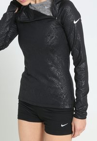 Mujer vestida con una camiseta deportiva de manga larga de mezcla de piel de serpiente negra y pantalones cortos negros de Nike, de pie contra un fondo claro y uniforme.