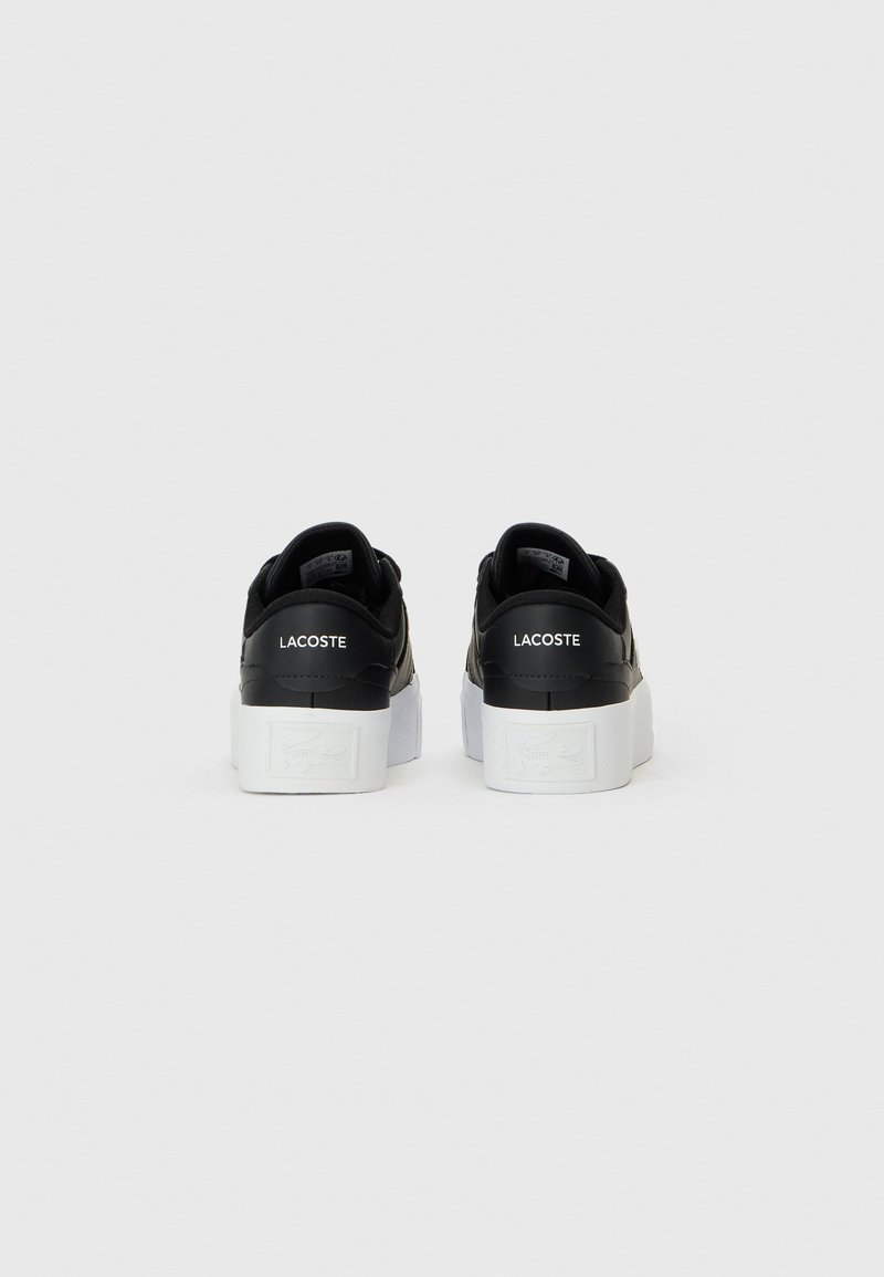 Scarpe in pelle nera con una suola bianca spessa, caratterizzate da un retro testurizzato e il nome del marchio "LACOSTE" stampato in bianco sul tallone.