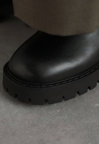 Black leather ankle boot featuring a rounded toe, thick ridged sole, and a smooth finish. The upper is sleek with subtle branding detail.