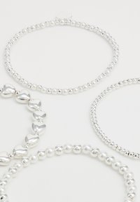 Štiri srebrne in bele zapestnice s perlicami, ena z metalnimi srčastimi perlicami, postavljene na enostavni beli ozadju.