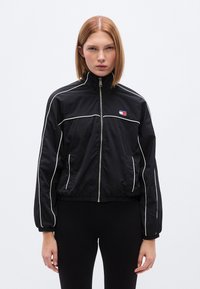 Μαύρο  μπουφάν με λευκή διακοσμητική ραφή, ψηλό γιακά, κλείσιμο με φερμουάρ και πλαϊνές τσέπες. Διαθέτει ένα μικρό λογότυπο στο αριστερό μέρος του στήθους.