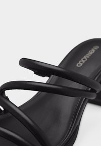 Μαύρο σανδάλι με τρεις λεπτές δερμάτινες λουρίδες πάνω από το πόδι και επίπεδη σόλα, εμφανίζοντας μέρος της εσωτερικής σόλας με τη σήμανση της μάρκας.