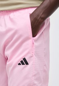 Χέρι ατόμου στην πλαϊνή τσέπη ροζ αθλητικού σορτς με μαύρο λογότυπο adidas και μικρές οπές αερισμού κατά μήκος της άκρης της τσέπης.