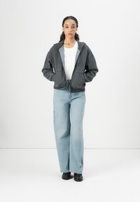 Gray zip-up hoodie with front pockets, worn over a white shirt. Light blue wide-leg jeans with a subtle graphic detail near the hem. Black shoes.