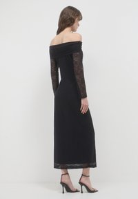 Robe noire décolletée avec des manches longues en dentelle, texture florale et coupe slim ; associée à des sandales à talons hauts noires.