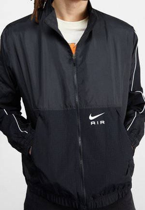 Schwarze Nike Air Zip-up-Jacke, getragen über einem weißen Hemd, mit Händen in den Taschen und weißen Paspeln an den Ärmeln.