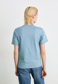 Personne avec des cheveux blonds courts portant un t-shirt bleu clair et un jean bleu, de dos devant un fond blanc uni.