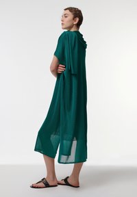 Vestido largo de tono azul verdoso, ligero y con capucha, que presenta mangas cortas y cortes laterales, combinado con sandalias planas negras.